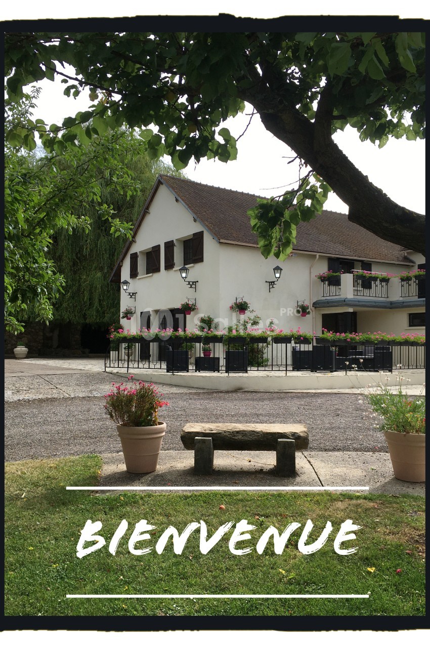 Maison traditionnelle avec volets en bois, entourée de fleurs, vue depuis un banc en pierre sous un arbre.