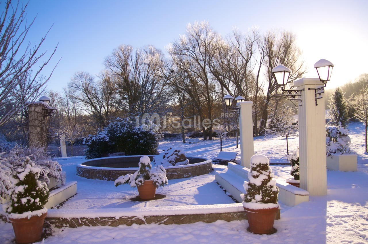 Jardin enneigé avec fontaine centrale, arbres dénudés et lampadaires sous un ciel bleu clair.