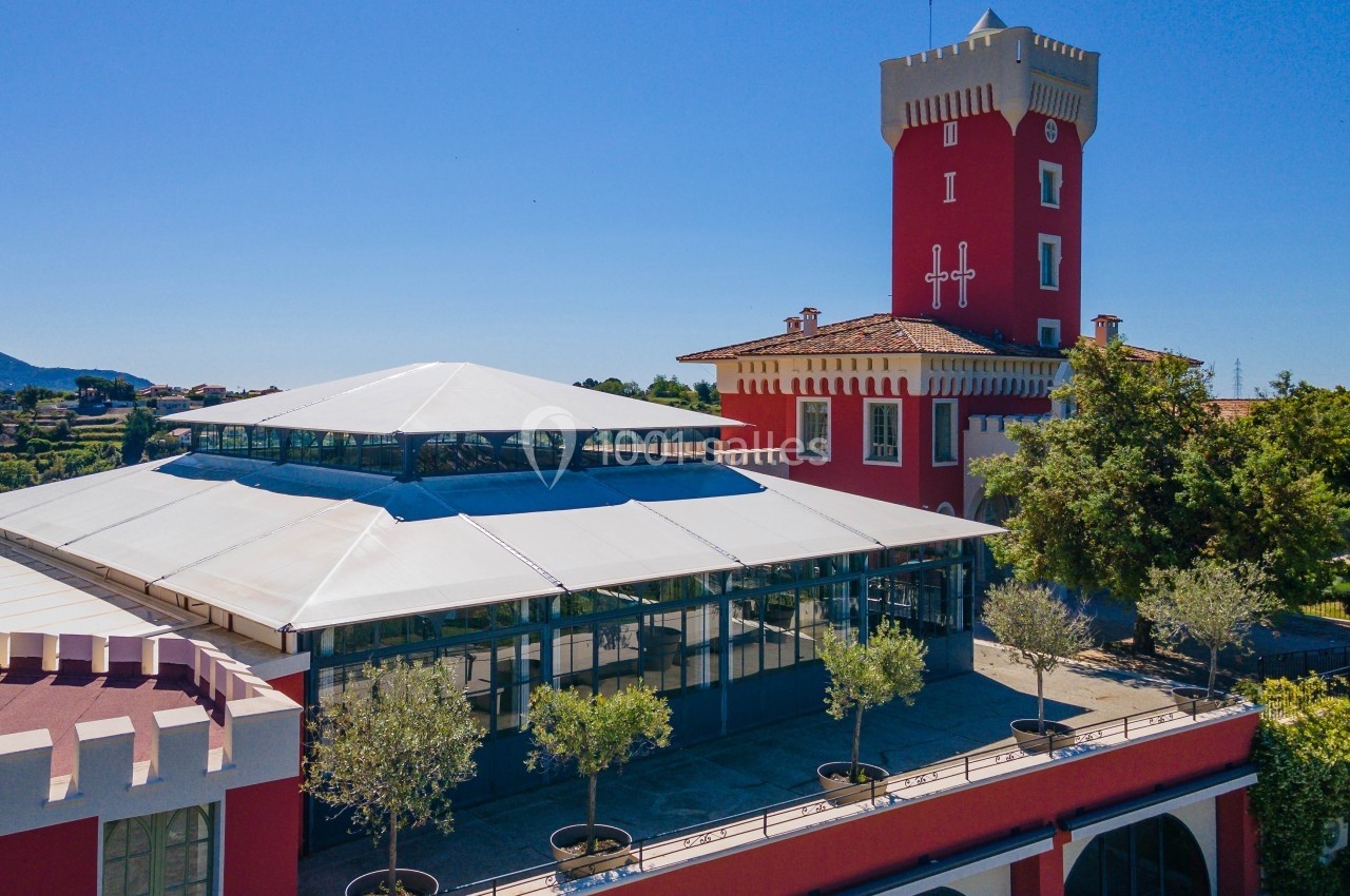 Bâtiment rouge avec une tour crénelée, une verrière moderne et des arbres en pots, sous un ciel bleu clair.