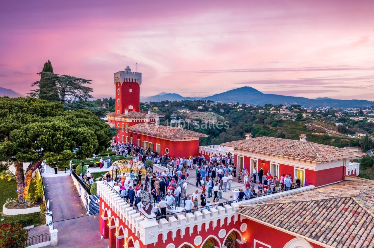 Vue aérienne d'un bâtiment rouge avec une terrasse animée, entouré de collines sous un ciel rose au coucher du soleil.