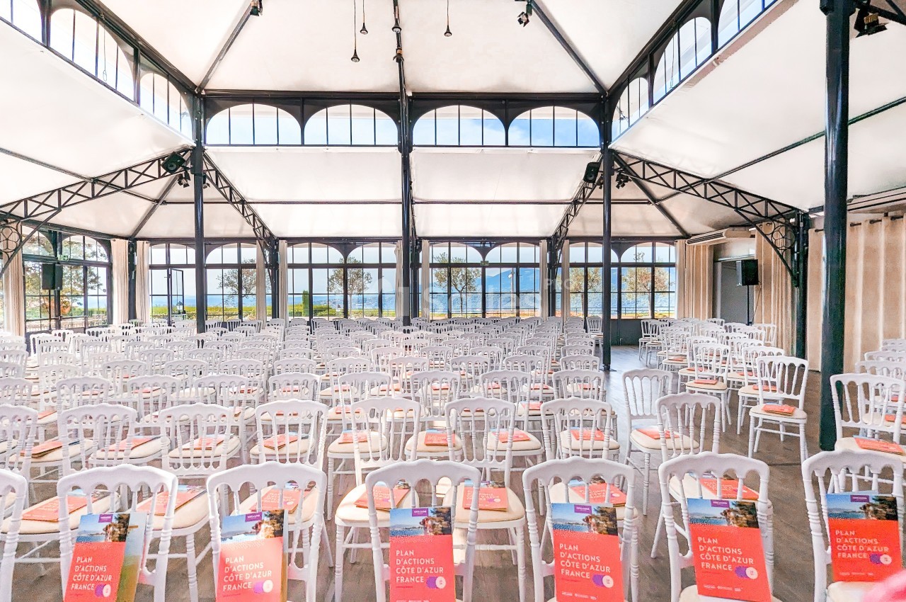 Salle lumineuse avec de grandes baies vitrées, remplie de chaises blanches alignées, chacune portant un livret rouge.