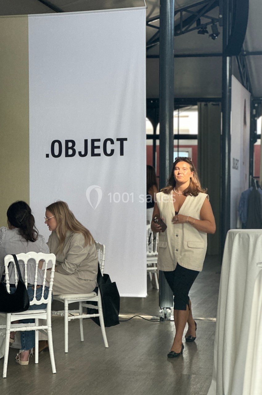 Une femme marche dans une salle lumineuse avec des tables occupées et une grande affiche marquée ’.OBJECT’.
