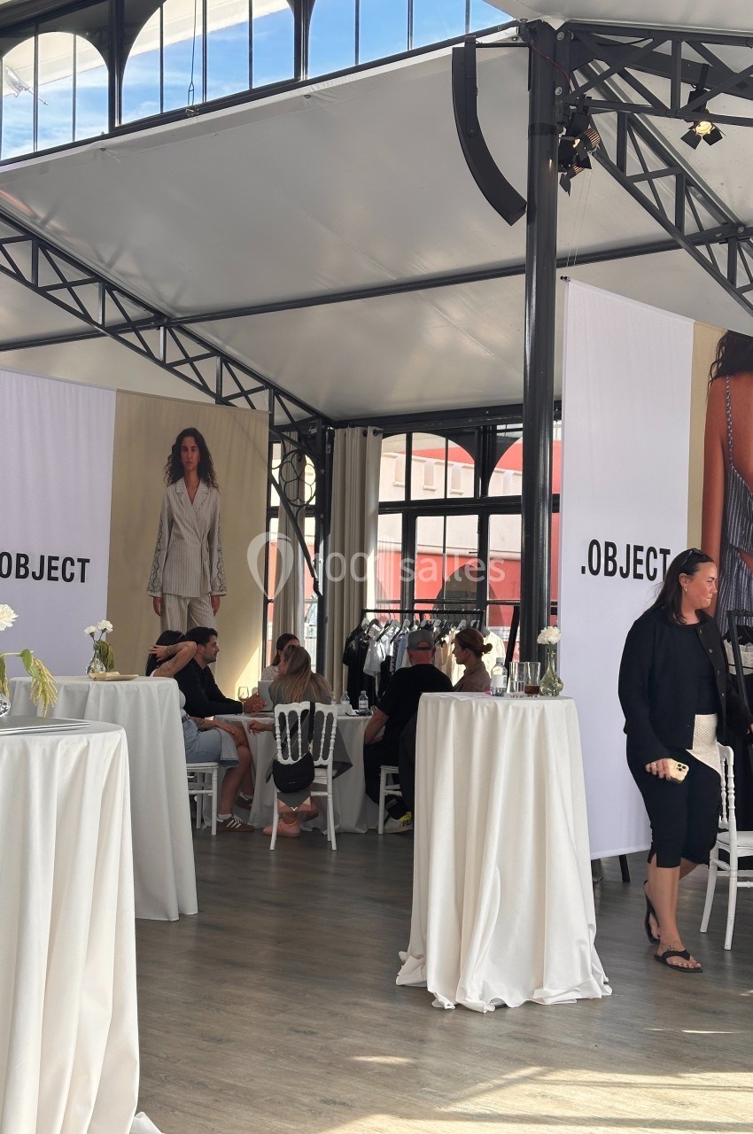 Espace lumineux avec tables hautes, affiches de mode et personnes discutant sous une structure métallique.