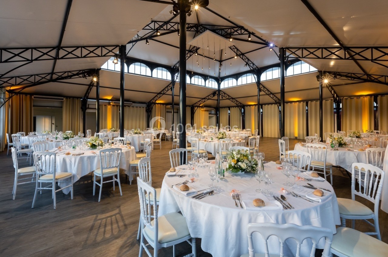 Salle de réception lumineuse avec tables rondes dressées, décorées de fleurs et entourées de chaises blanches.