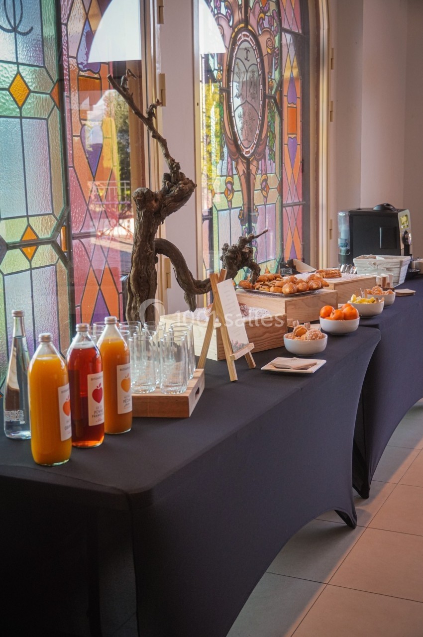 Buffet avec jus de fruits, viennoiseries, fruits frais et café, disposé sur une table devant des vitraux colorés.