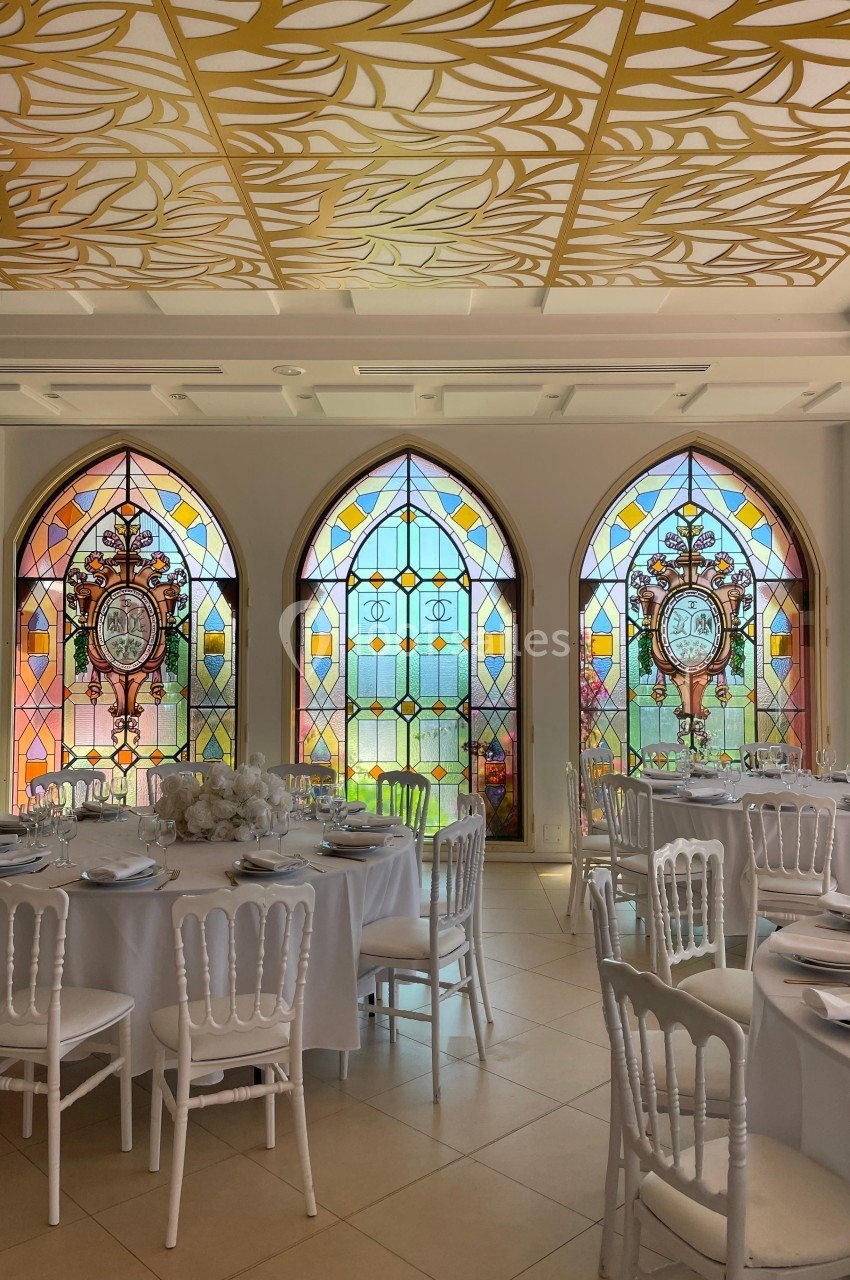 Salle de réception lumineuse avec tables dressées et grandes fenêtres ornées de vitraux colorés.