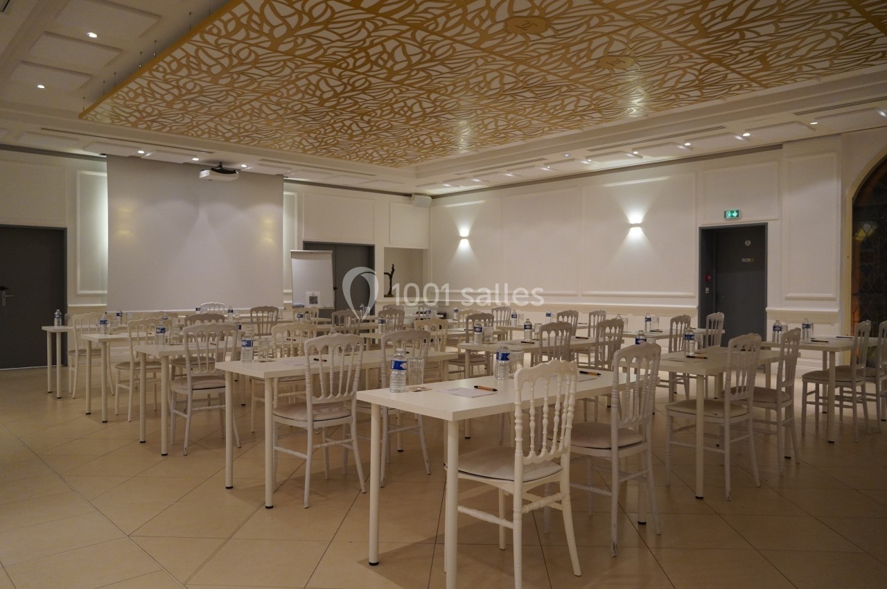 Salle de réunion lumineuse avec des tables blanches alignées, des chaises et un plafond décoré de motifs dorés.