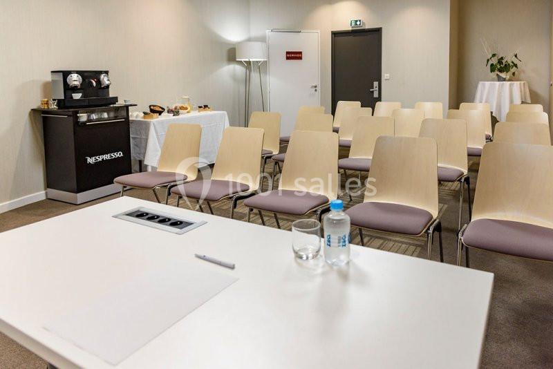 Salle de réunion avec chaises alignées, table équipée de prises électriques et boissons, coin café en arrière-plan.