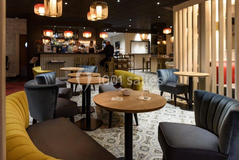 Espace lounge avec tables en bois, fauteuils colorés et éclairage tamisé, bar en arrière-plan.