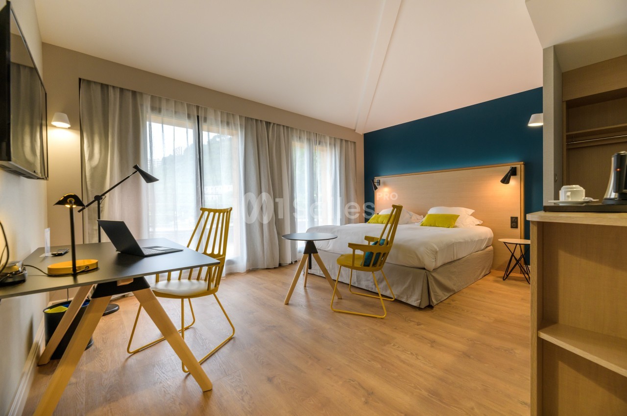 Chambre lumineuse avec lit double, bureau équipé d'un ordinateur portable, chaises jaunes et vue sur l'extérieur.