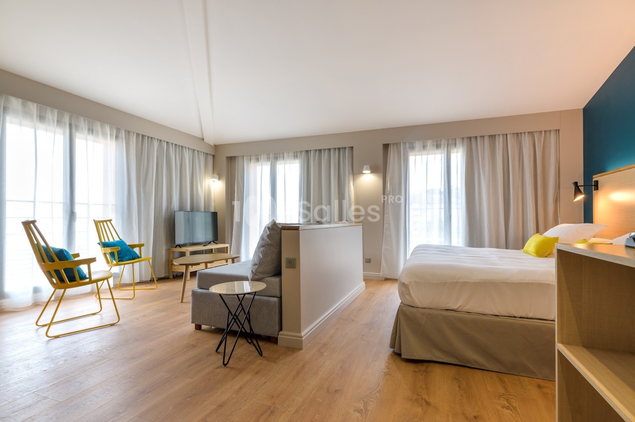 Chambre lumineuse avec lit double, coin salon, fauteuils jaunes, téléviseur et grandes fenêtres avec rideaux.