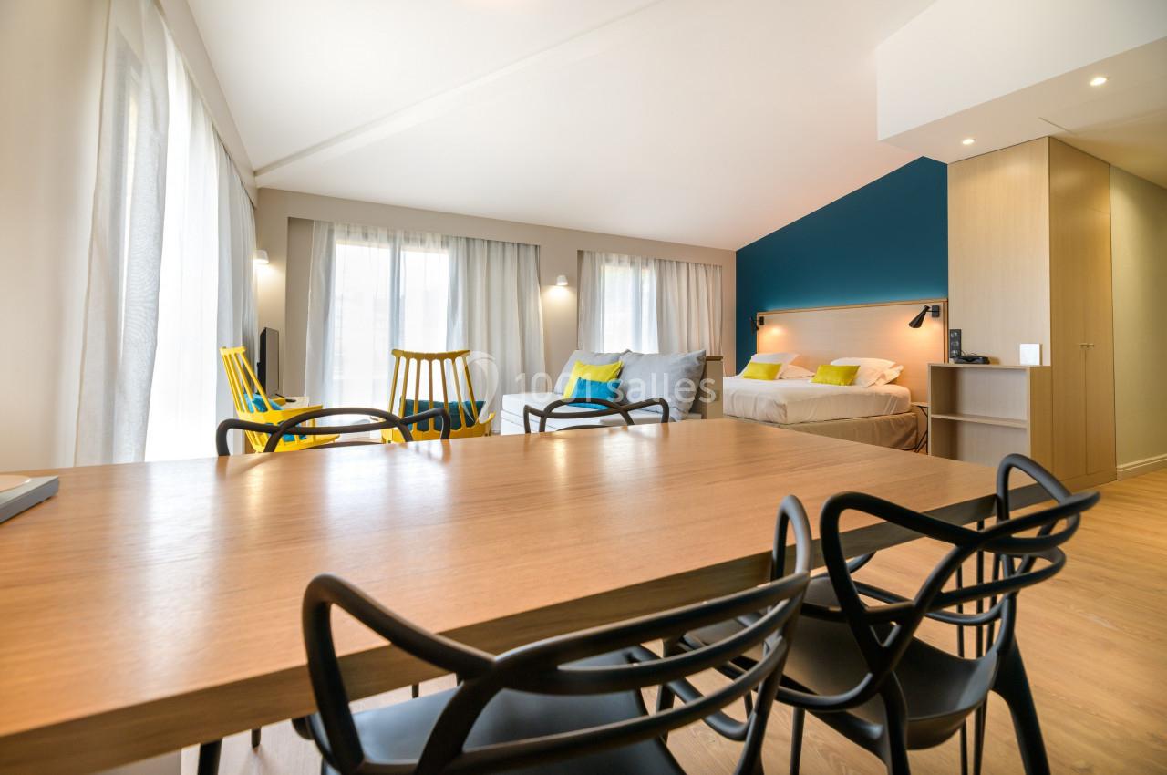 Salle lumineuse avec table en bois, chaises modernes, coin salon, lit double et murs aux tons neutres et bleu.