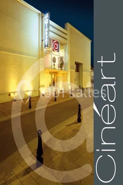 Façade d'un cinéma éclairé de nuit, avec une entrée vitrée et des bornes noires le long du trottoir.