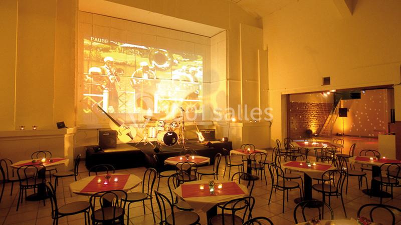 Salle de spectacle avec tables et chaises, éclairage tamisé, scène équipée d'une batterie et projection murale.