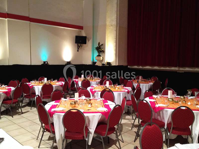 Salle de réception avec tables rondes dressées, nappes blanches et roses, chaises rouges, éclairage tamisé.