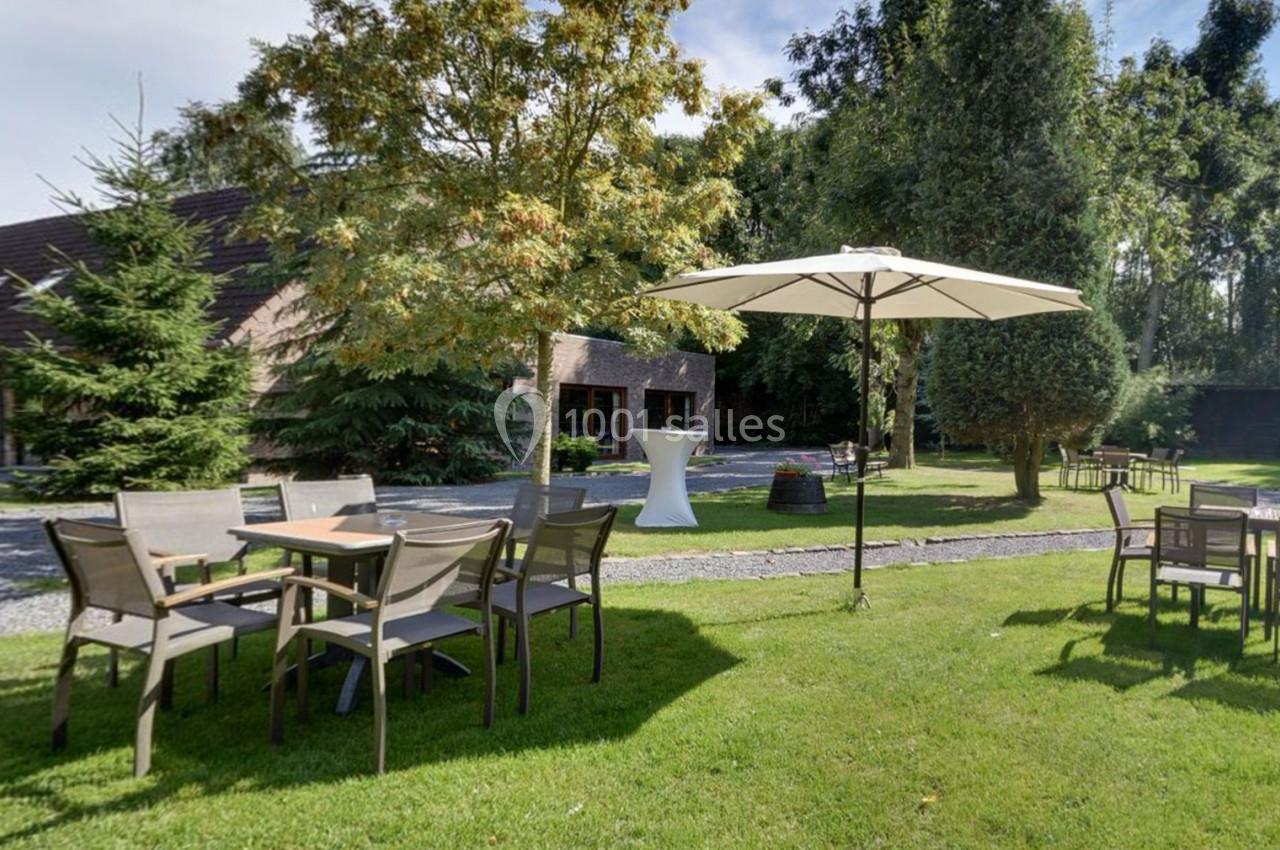 Location salle Mouscron (Hainaut) - Le Belvédère #22 Tables et chaises en plein air sur une pelouse, avec un parasol et des arbres en arrière-plan.