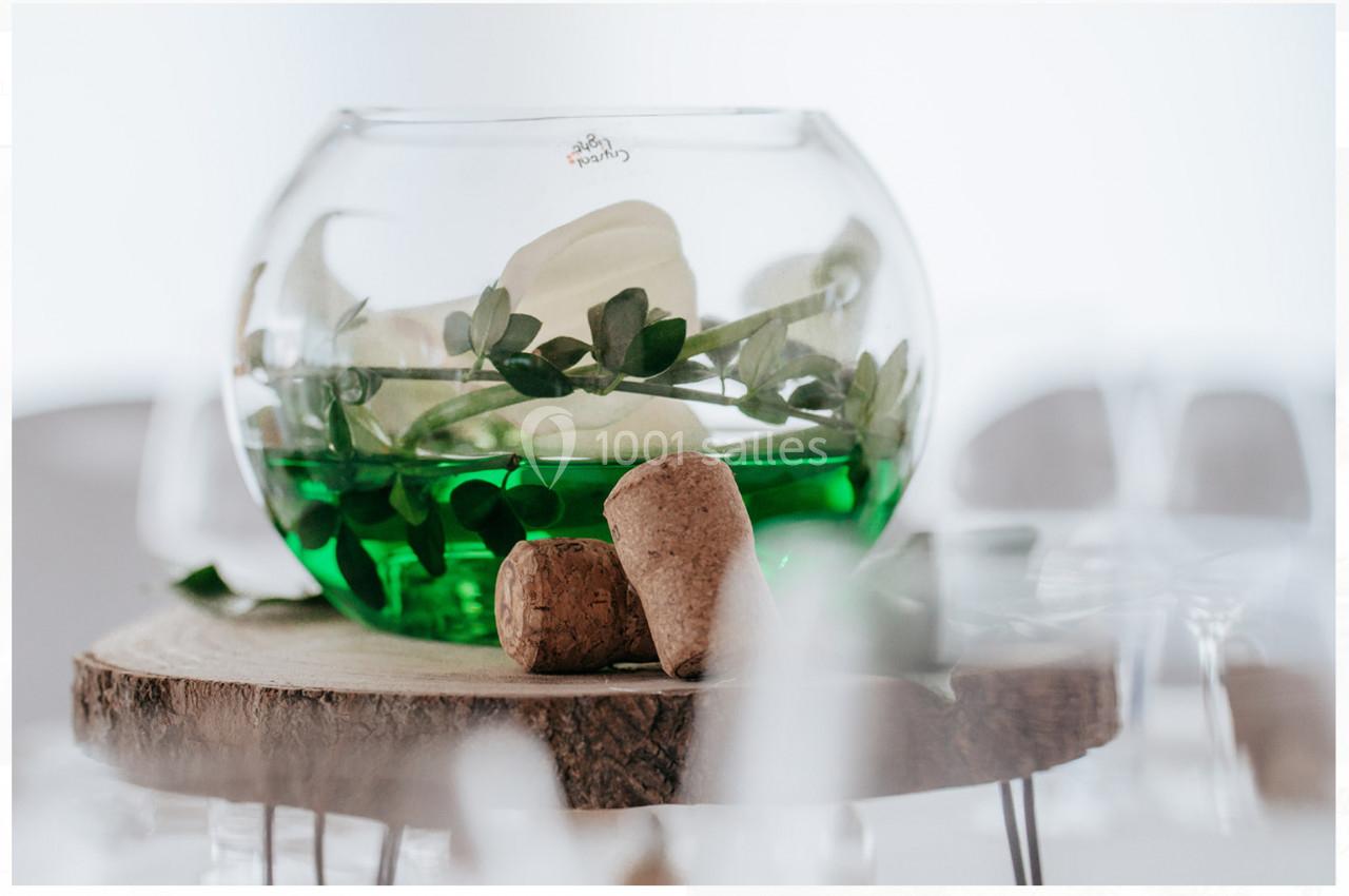 Location salle Mouscron (Hainaut) - Le Belvédère #9 Vase rond avec liquide vert, feuillage et fleurs blanches, posé sur une tranche de bois avec deux bouchons en liège.
