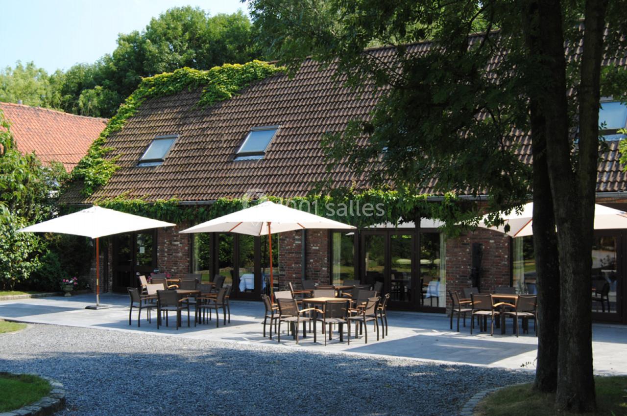 Location salle Mouscron (Hainaut) - Le Belvédère #3 Terrasse extérieure avec tables, chaises et parasols devant un bâtiment en briques entouré de verdure.
