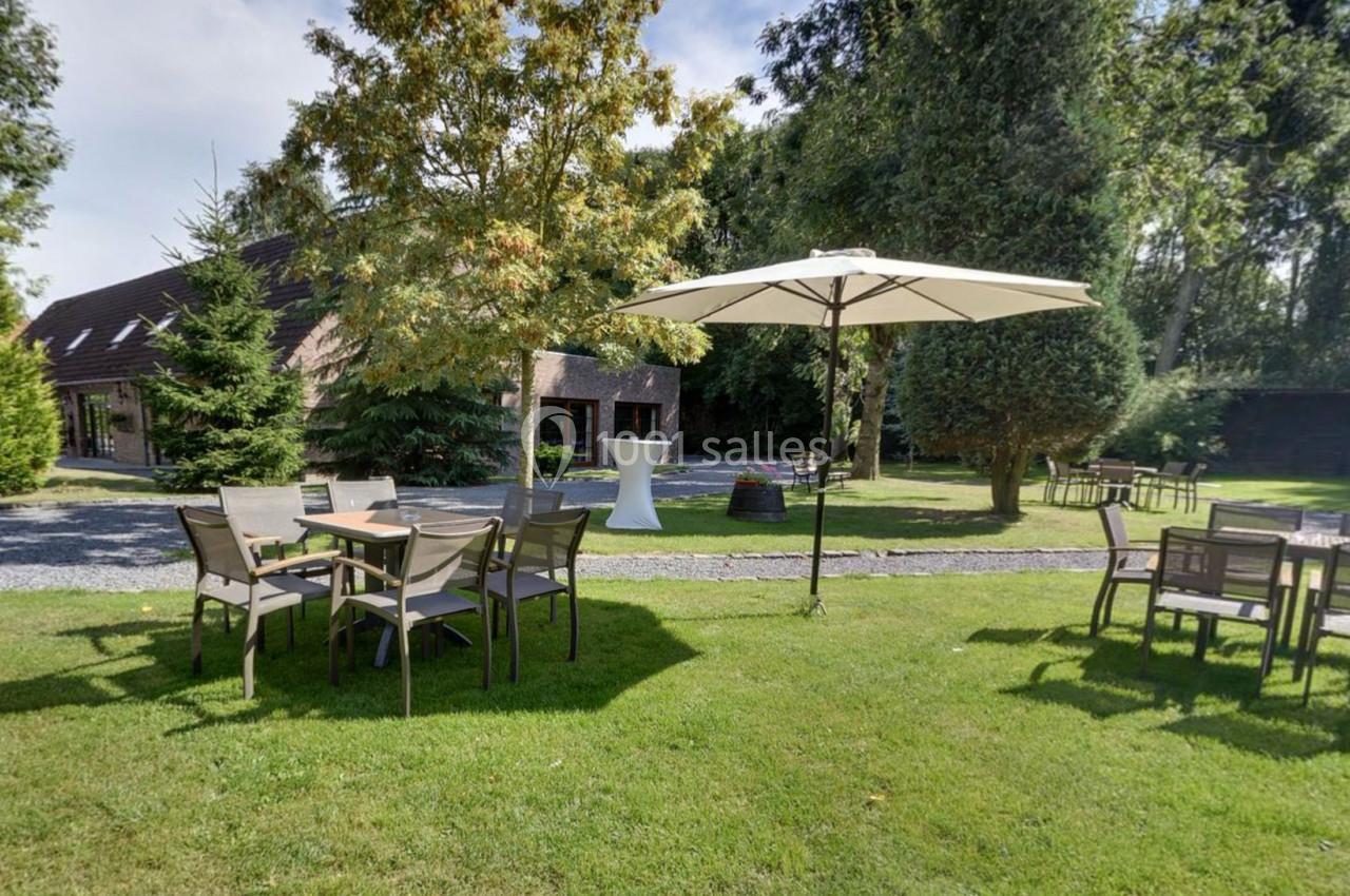 Location salle Mouscron (Hainaut) - Le Belvédère #6 Jardin avec tables, chaises et parasol sur pelouse, entouré d'arbres et d'une maison en arrière-plan.