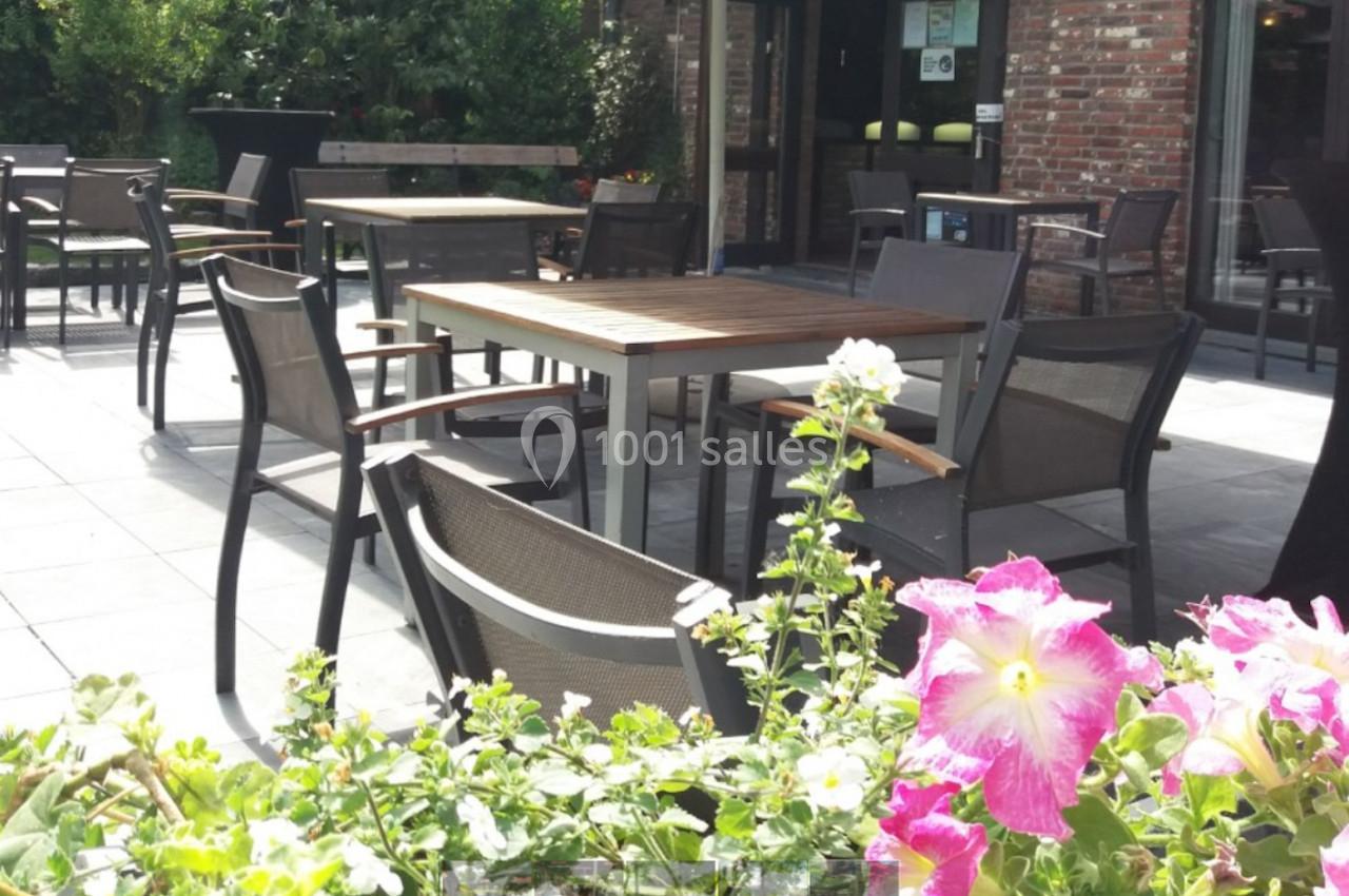 Location salle Mouscron (Hainaut) - Le Belvédère #18 Terrasse extérieure avec tables et chaises en bois et métal, entourée de verdure et de fleurs colorées au premier plan.