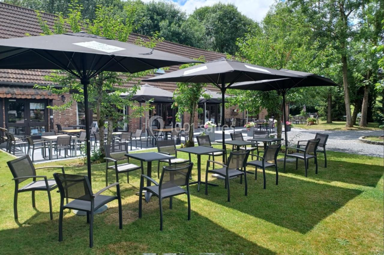 Location salle Mouscron (Hainaut) - Le Belvédère #20 Terrasse extérieure avec tables, chaises et parasols noirs, entourée de verdure et bâtiments en briques.