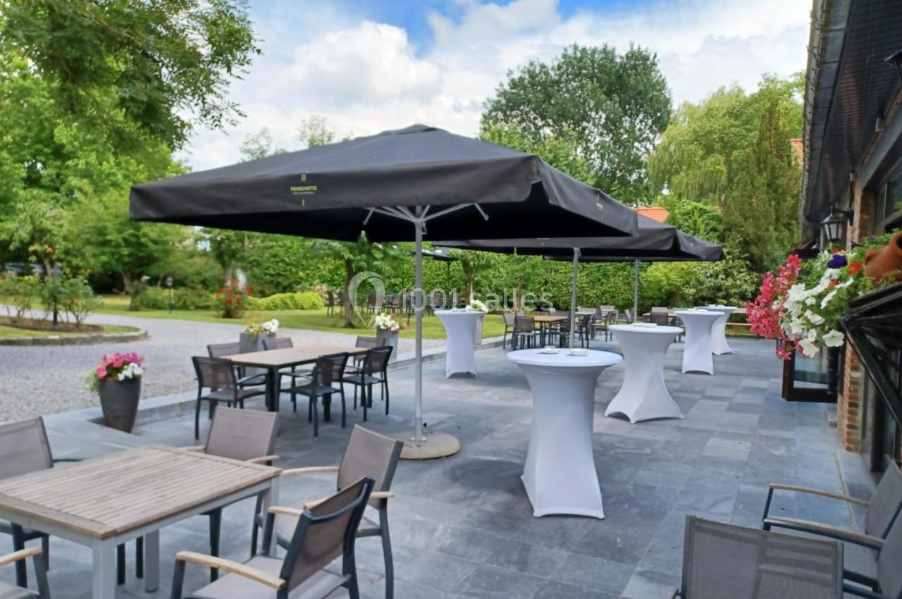 Location salle Mouscron (Hainaut) - Le Belvédère #21 Terrasse extérieure avec tables, chaises, parasols noirs et tables hautes, entourée de verdure et fleurs.