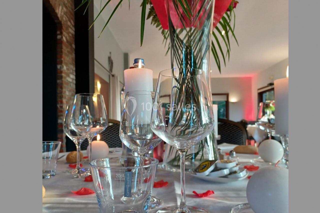 Location salle Mouscron (Hainaut) - Le Belvédère #14 Table dressée avec des verres, bougies et pétales rouges, décorée d'un grand vase avec des feuilles et fleurs.