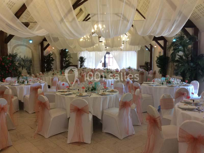 Salle de réception décorée avec des tables rondes, nappes blanches, chaises ornées de nœuds orange et voilages suspendus.