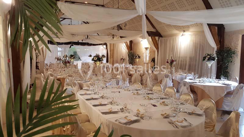 Salle de réception décorée avec des tables rondes dressées, chaises couvertes de housses et voilages suspendus.