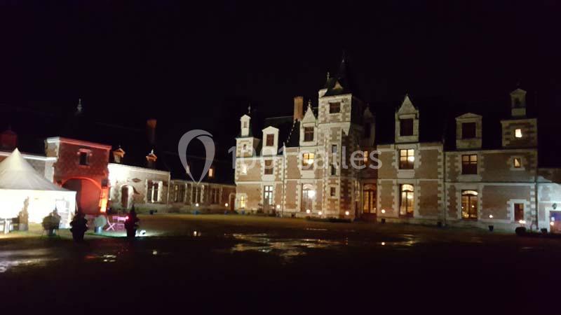 Façade d'un château illuminé la nuit, avec des fenêtres éclairées et une tente blanche sur la gauche.