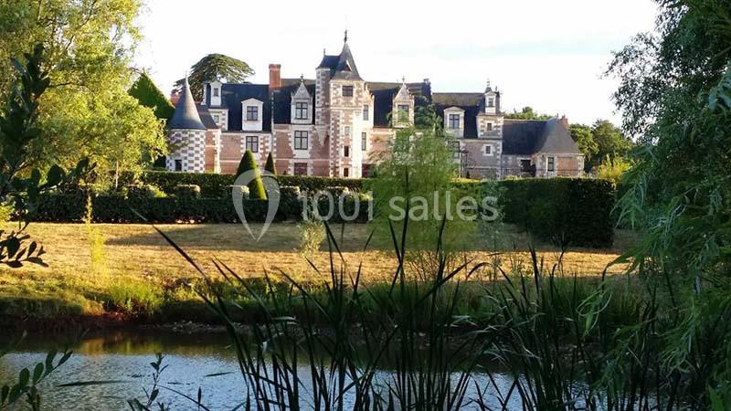 Vue d'un château en pierre entouré de verdure, avec un étang au premier plan et un ciel dégagé.