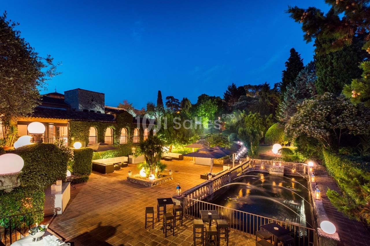 Terrasse éclairée en soirée avec tables, bassin d'eau et végétation luxuriante entourant l'espace.