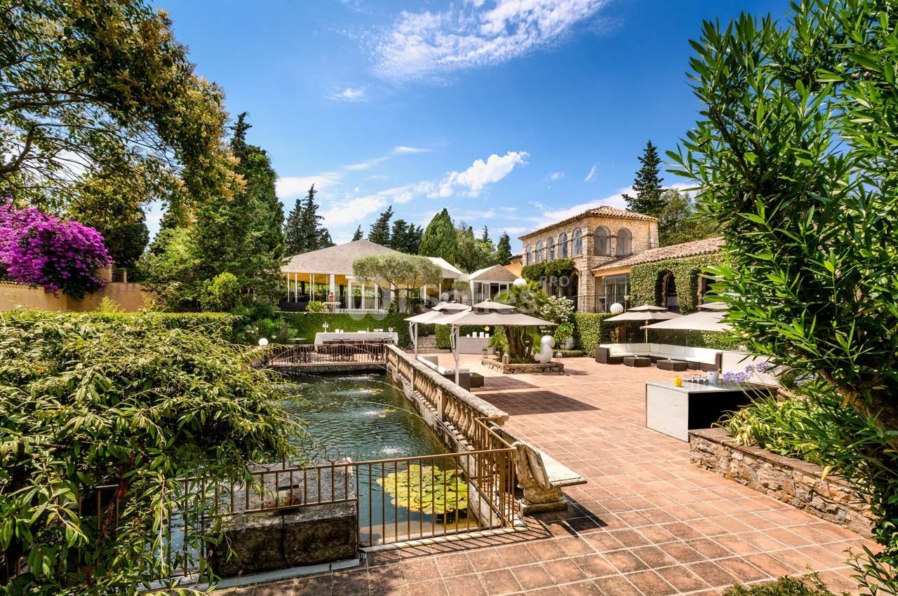 Jardin avec étang, terrasse en pierre, parasols et bâtiment en pierre sous un ciel bleu ensoleillé.