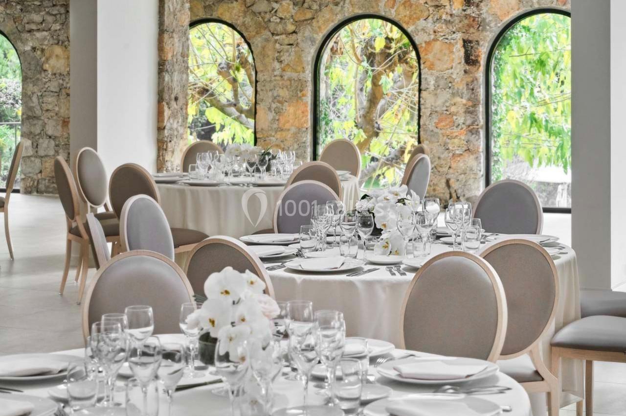 Salle élégante avec tables rondes dressées, nappes blanches, chaises grises et grandes fenêtres en arc donnant sur un jardin.