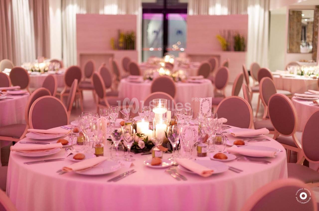 Salle de réception décorée avec des tables rondes, nappes roses, bougies et vaisselle élégante.