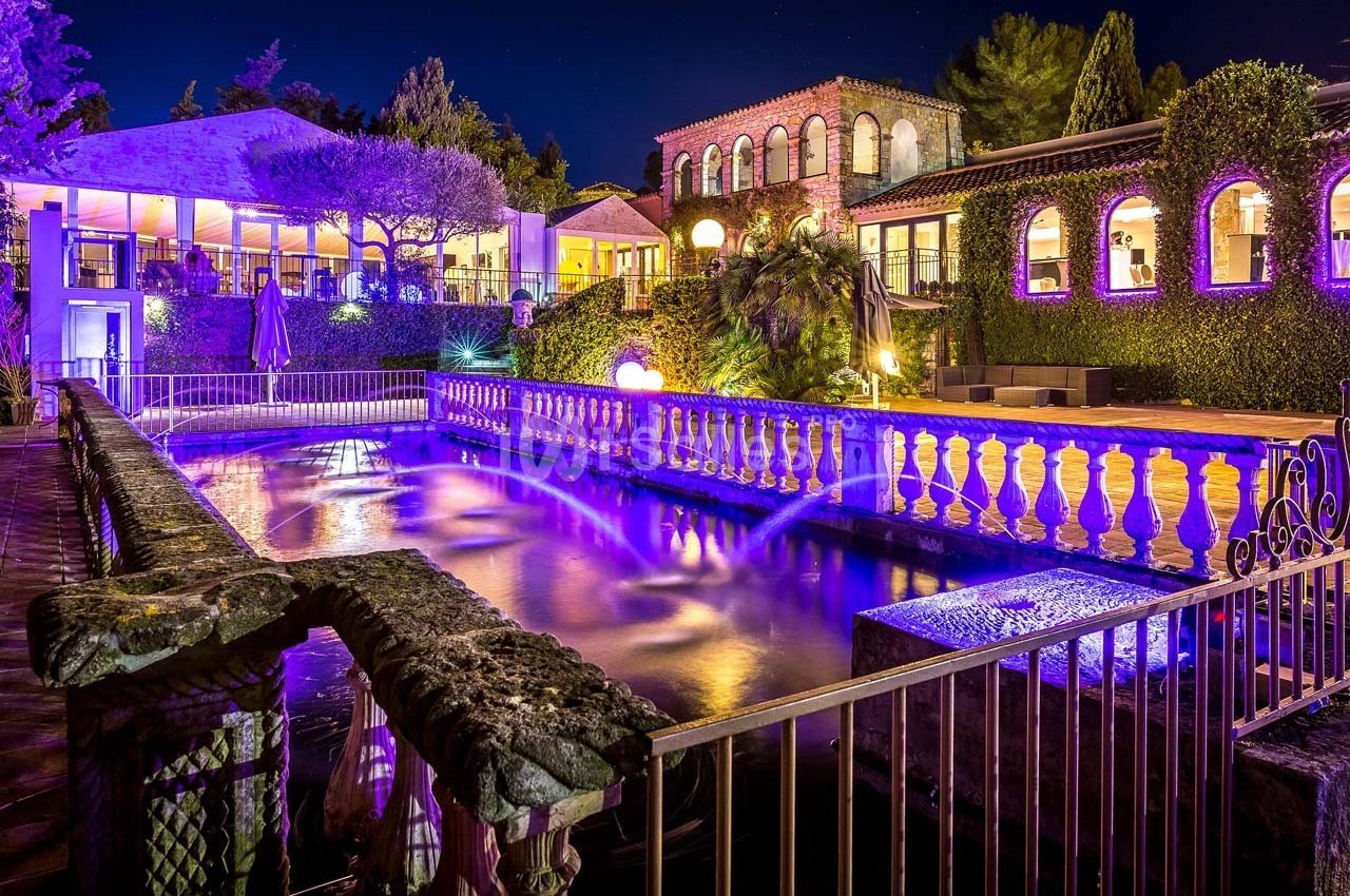 Vue nocturne d'un domaine avec piscine éclairée en violet, entourée de bâtiments et de végétation.