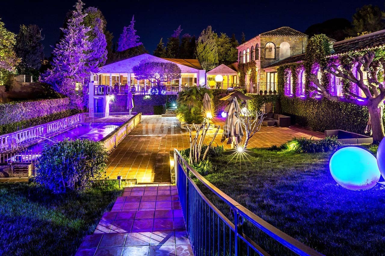 Jardin éclairé de nuit avec piscine, terrasse en pavés et bâtiments entourés de végétation.