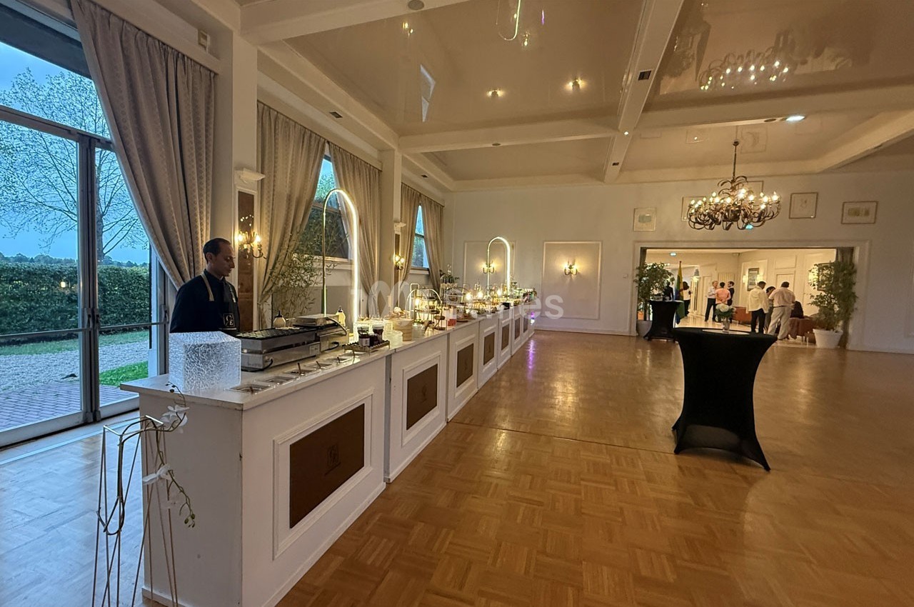 Salle de réception élégante avec un buffet éclairé, des lustres suspendus et de grandes fenêtres donnant sur un jardin.