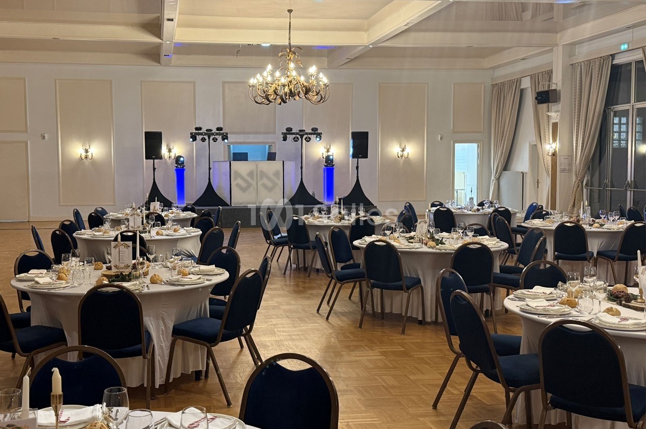 Salle de réception élégante avec tables rondes dressées, chaises bleues et espace pour un DJ en fond.