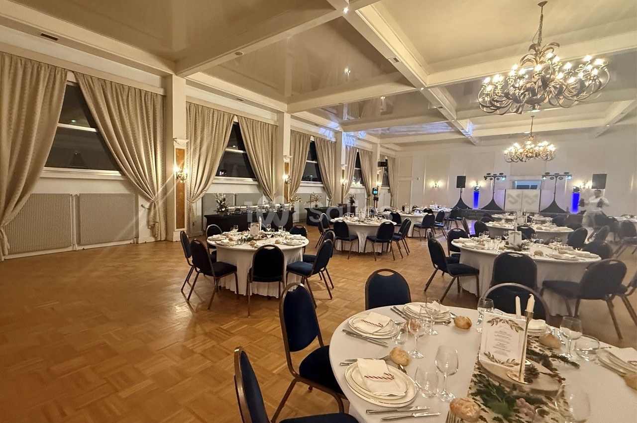 Salle de réception élégante avec tables rondes dressées, chandeliers suspendus et rideaux beiges aux fenêtres.
