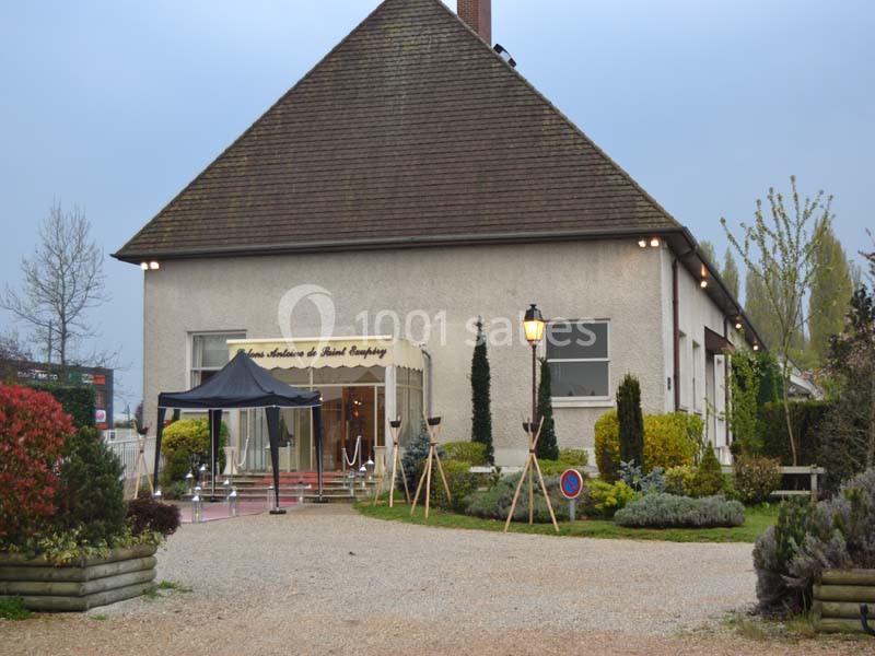Bâtiment avec une entrée vitrée, un auvent noir et un chemin gravillonné entouré de végétation.