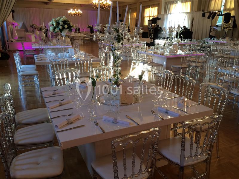 Salle de réception élégante avec tables décorées de fleurs blanches, chaises transparentes et éclairage chaleureux.