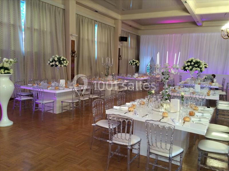 Salle de réception décorée avec des tables dressées, des chaises transparentes et des compositions florales blanches.