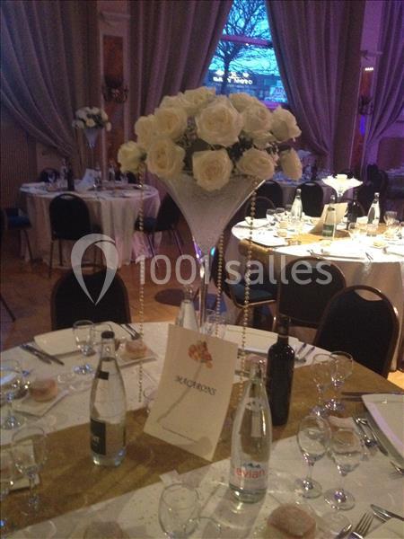 Centre de table avec des roses blanches dans un vase en verre, entouré de tables dressées pour un événement.