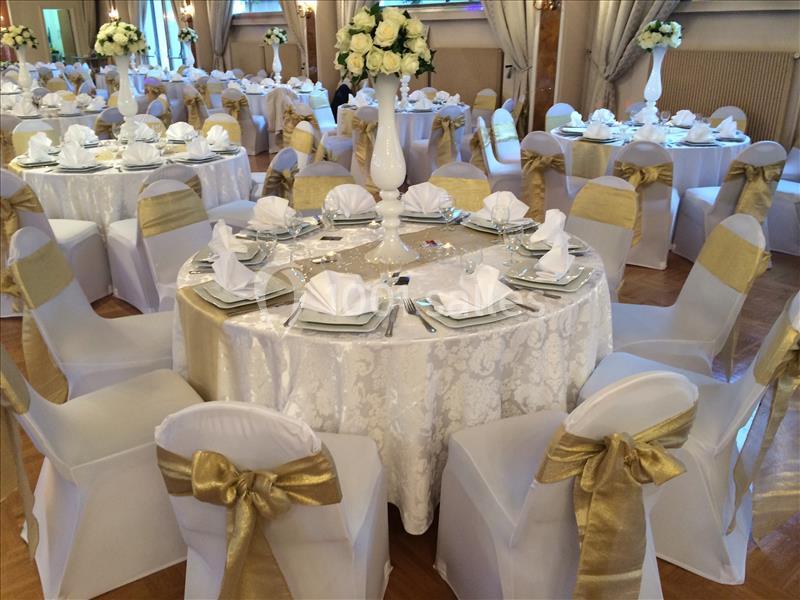 Salle de réception décorée avec des tables rondes, nappes blanches, chaises ornées de nœuds dorés et centres de table…