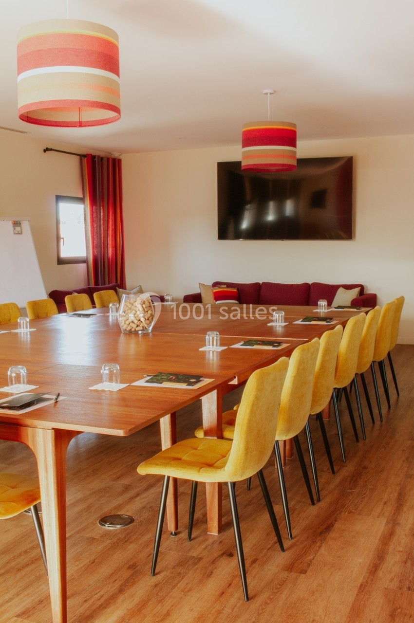 Salle de réunion lumineuse avec une grande table en bois, des chaises jaunes et un écran mural.