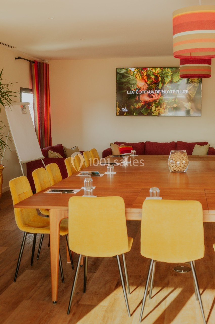 Salle de réunion lumineuse avec table en bois, chaises jaunes, écran mural et tableau blanc.
