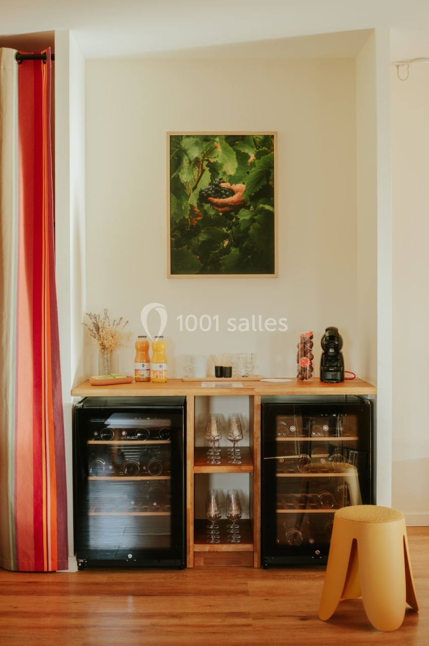 Coin bar avec deux caves à vin, une machine à café, des bouteilles de jus et un tabouret jaune devant un mur décoré.