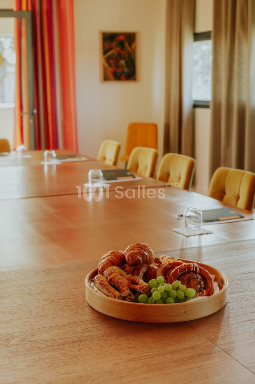 Assiette de viennoiseries et raisins posée sur une grande table en bois dans une salle lumineuse avec des chaises jaunes.