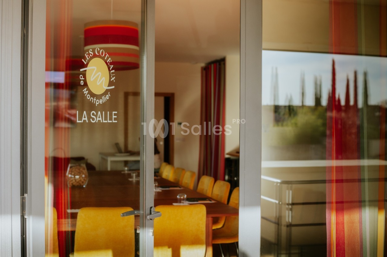 Salle de réunion lumineuse avec table en bois, chaises jaunes et rideaux rouges, vue à travers une porte vitrée.